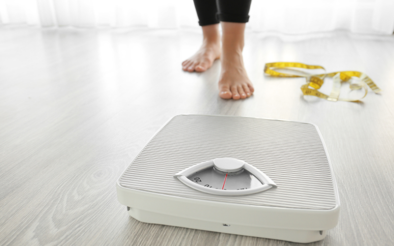 Pessoa se aproximando de balança com fita métrica no chão, simbolizando o desafio do emagrecimento.