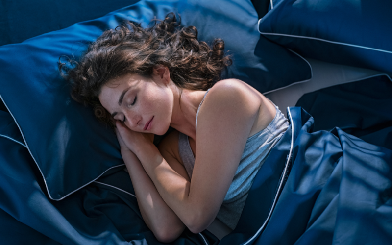 Mulher deitada em cama azul escura, dormindo profundamente com expressão serena.