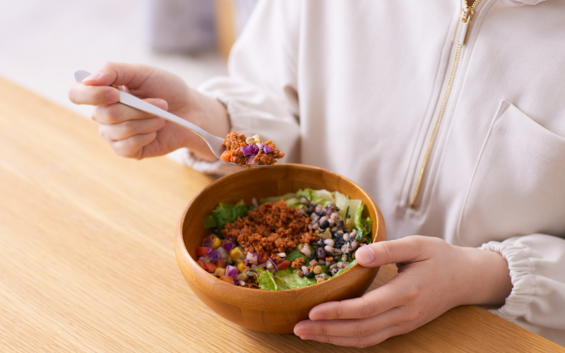 Mão segurando colher com salada colorida de feijão, vegetais e proteína vegetal triturada, dentro de uma tigela de madeira.