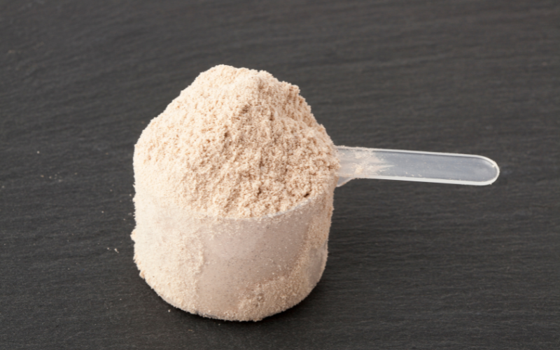 Scoop transparente cheio de whey protein em pó sobre superfície escura.